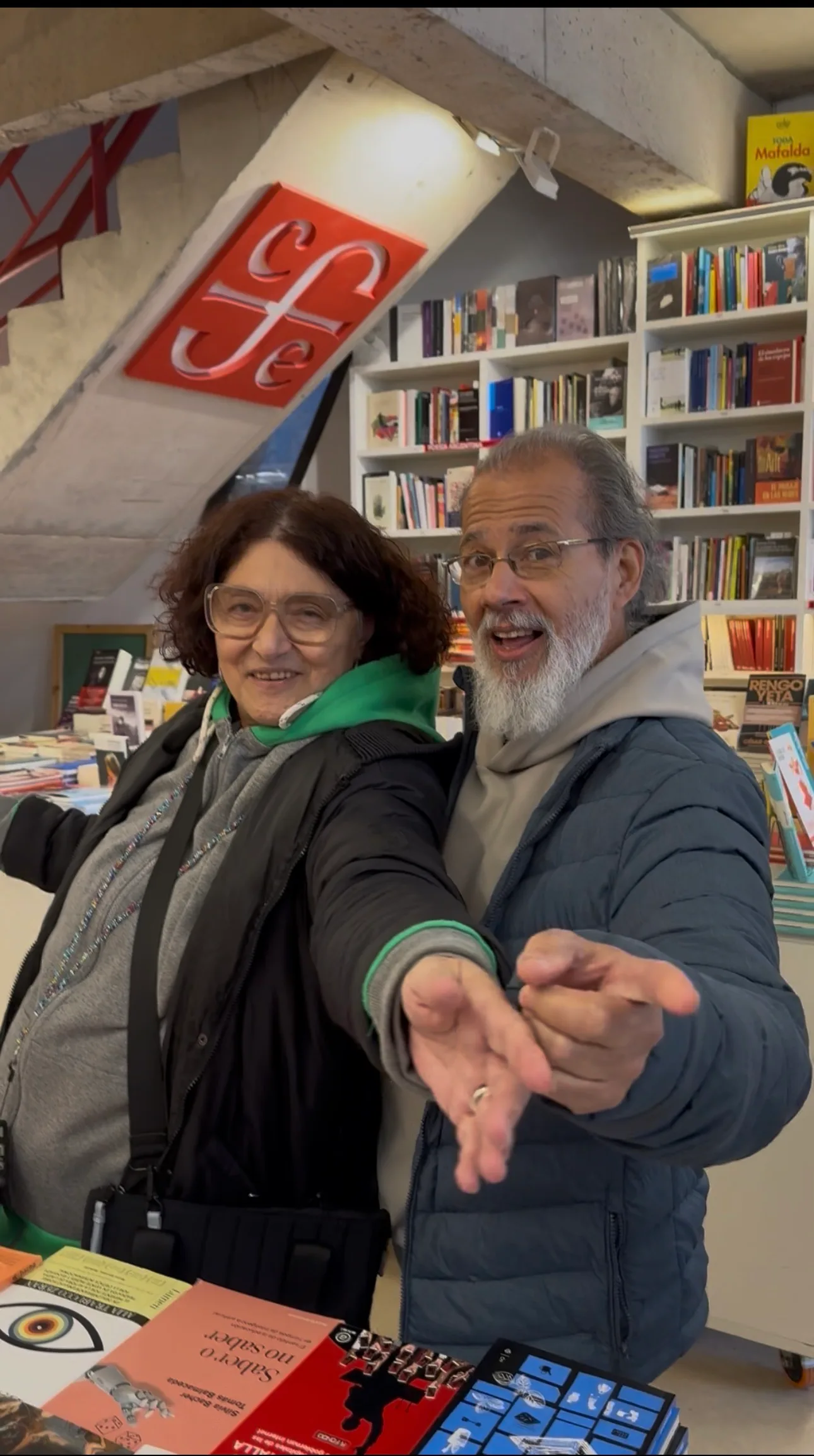 Alejandra Giménez y Fernando Rivero en una librería, entre mesas de libros, señalando hacia la cámara